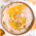 Creamy white bean dip in a bowl, served with a drizzle of olive oil, herbs and baked tortilla chips