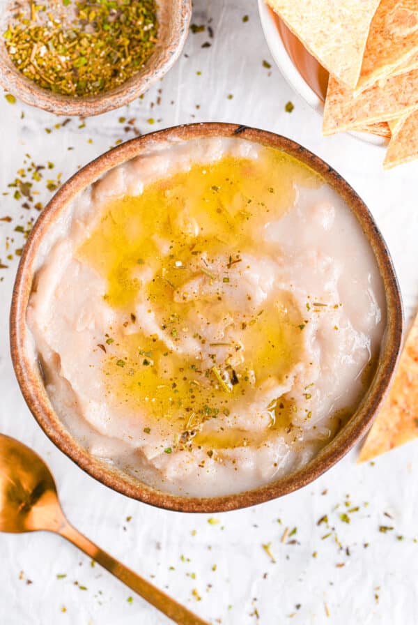 Creamy white bean dip in a bowl, served with a drizzle of olive oil, herbs and baked tortilla chips