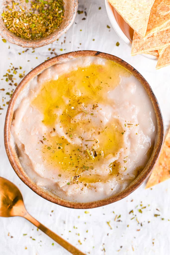 Creamy white bean dip in a bowl, served with a drizzle of olive oil, herbs and baked tortilla chips